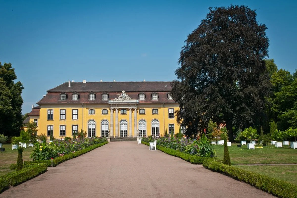 Schloss Mosigkau Ein Schlossgarten erstreckt sich rechts und links neben einem Schloss. Ein breiter Weg führt zu dem großen, gelben Schlossgebäude.